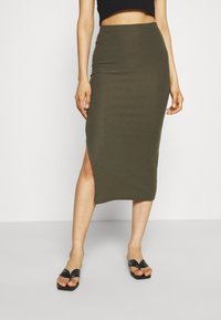 Žena v olivově zelené ribbed midi sukni s bočním rozparkem, černém cropped topu a černých sandálech na podpatku s čtvercovou špičkou, stojící na bílém podlaze.