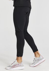 Svarta tajta leggings av mjuk tyg med dragkedja vid ankeln, tillsammans med grå och vita träningsskor med röda accenter.