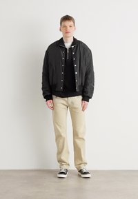 Jaqueta bomber preta com fecho de zíper e pressão sobre um suéter preto. Calças bege e sapatilhas pretas completam o visual, contra um fundo neutro.