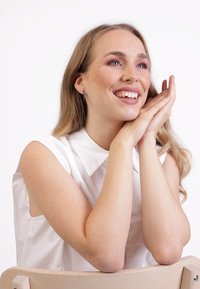 Eine Frau mit langen Haaren, die ein ärmelloses weißes Hemd mit Kragen trägt, lehnt lächelnd mit den Händen an ihrem Gesicht auf einem hellen Holzstuhl.