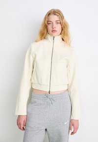 Kreminės spalvos zip-up striukė su aukštu apykakle, briaunota apačia ir Nike logotipu; derinama su pilkomis treniruotėmis, turinčiomis raištelio juosmenyje.