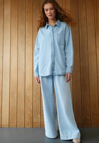 Lichtblauwe denimshirt met een zak en knoopsluiting, gecombineerd met wijdvallende lichtblauwe denimbroek. Gladde textuur, relaxte pasvorm.