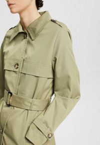 Khaki trencz ma zapiętą kołnierz, przewiązany w talii pas i dwie kieszenie na piersi. Wykonany z gładkiego materiału, z widocznymi szwami i akcentami guzików.