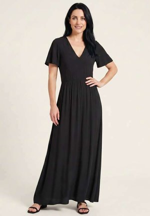 Femme aux longs cheveux noirs portant une robe longue noire à manches courtes et encolure en V, ainsi que des sandales noires, debout, une main sur la hanche.