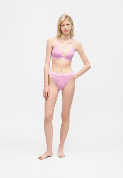 Γυναίκα που στέκεται ξυπόλυτη φοράει ασορτί ανοιχτόροζ σουτιέν και σλιπ με ψηλή μέση και λογότυπο στη ζώνη, μπροστά σε απλό λευκό φόντο.