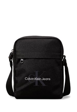 Bandolera negra hecha de tejido texturizado. Cuenta con un bolsillo frontal con cremallera y el logo "Calvin Klein Jeans" en blanco y gris.