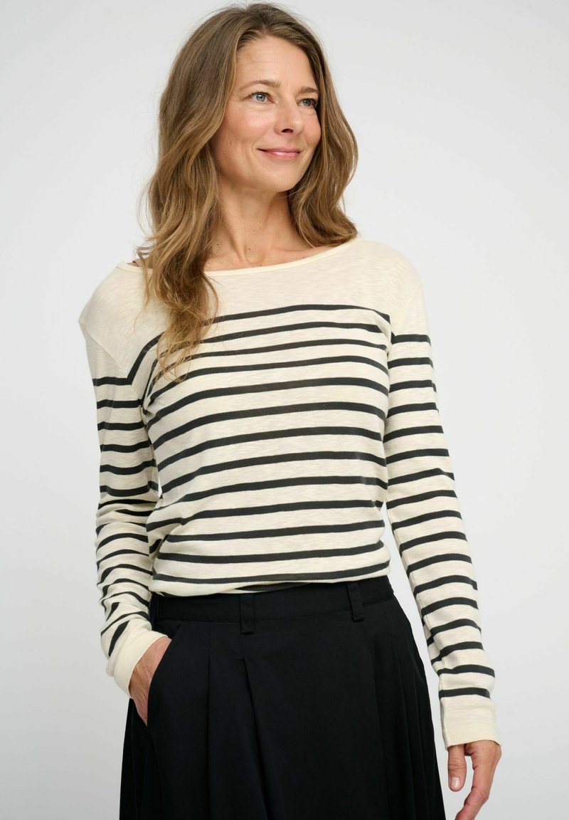 Striped long-sleeve top in cream with black horizontal stripes, featuring a relaxed fit, wide neckline, and ribbed cuffs. Paired with black trousers.