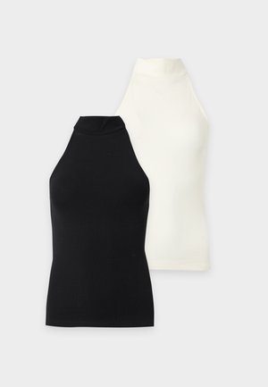 Femme debout sur un fond blanc portant un haut noir à col halter sans manches, une jupe midi vert olive, des chaussettes blanches et des baskets blanches.