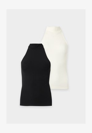 Femme debout sur un fond blanc portant un haut noir à col halter sans manches, une jupe midi vert olive, des chaussettes blanches et des baskets blanches.