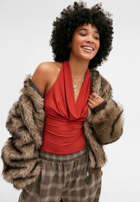Top rojo con cuello halter drapeado en el frente, combinado con una chaqueta de pelaje sintético marrón y pantalones de patrón de cuadros con cintura elástica.