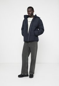 Puffermantel in Navyblau, mit Reißverschluss vorne und Kapuze. Getragen über einem leichten Hemd. Kombiniert mit grauen, weit geschnittenen Jeans und schwarzem Schuhwerk.