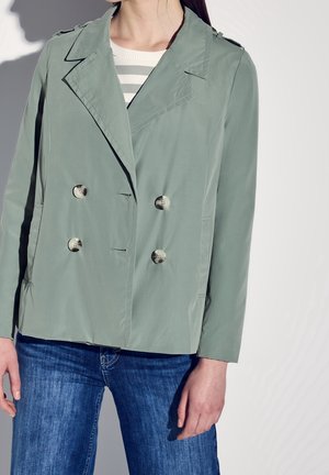 Kvinde iført lys grøn dobbeltradet jakke over en hvid og grå stribet skjorte med blå jeans mod en ensfarvet baggrund.