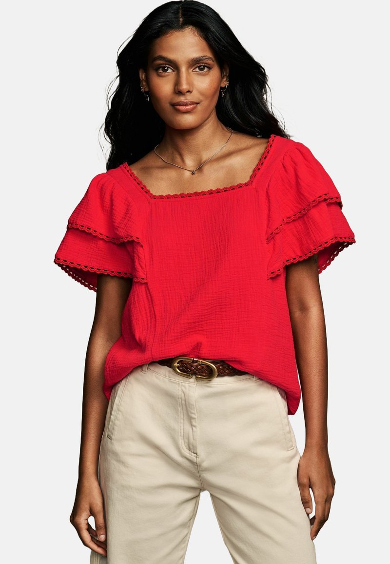 Top rosso a maniche a sbuffo con una finitura testurizzata, scollatura quadrata e orlo ondulato, abbinato a pantaloni beige e una cintura intrecciata.