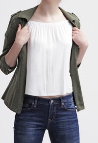 Giacca militare verde oliva indossata sopra una blusa bianca plissettata senza spalline, abbinata a jeans blu scuro, con attenzione alla trama e alla vestibilità.