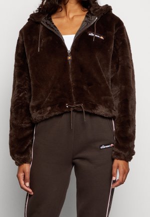 Frau trägt eine dunkelbraune Kapuzenjacke aus Kunstpelz mit Reißverschluss und passende Jogginghose, beide mit ellesse-Logos auf der Brust und dem Oberschenkel.