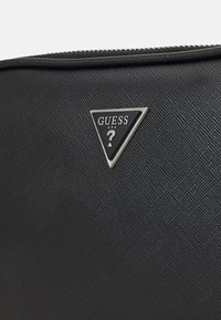 Borsa a tracolla in tessuto nero con texture, caratterizzata da un emblema triangolare in argento con la scritta "GUESS" e un punto interrogativo. Chiusura con cerniera nella parte superiore.