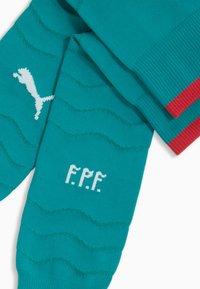 Calzini sportivi turchesi con motivi a onde, logo Puma bianco e scritta "F.P.F." in bianco sulla parte del piede. Accenti rossi sulla parte superiore.