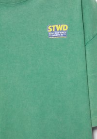 T-shirt en tissu vert avec un logo texte jaune et blanc indiquant ""STWD Stuff This World Delights In"" sur le haut de la poitrine.