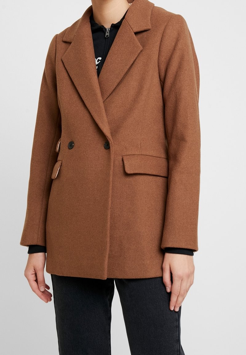 Mujer vistiendo un abrigo de lana marrón de doble botonadura con botones negros, sobre una parte superior negra con cremallera y pantalones negros, de pie contra un fondo liso.