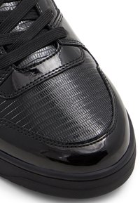 Black sneakers with a glossy patent finish, featuring a textured pattern and black laces. The sole is rubber and slightly rounded.
