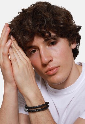 Black braided leather bracelet on wrist. Model has tousled dark hair and is wearing a plain white t-shirt, hands resting near face.