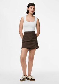 Donna con capelli corti e scuri che indossa un top bianco senza maniche, una skort a righe marroni e scarpe da ginnastica beige e nere, in piedi con le mani dietro la schiena.