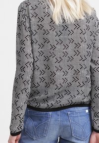 Pull en noir et blanc à motifs avec un col rond et des manches longues, présentant un ourlet noir uni, porté sur un jean bleu.
