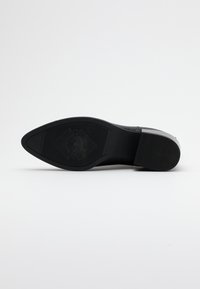 Suela de zapato negra con forma puntiaguda. Fabricada en goma, presenta un acabado texturizado y un detalle de logo en relieve en el centro.