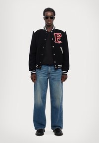 Zwarte varsityjack met witte accenten en rode geborduurde letter "F," gecombineerd met blauwe wijde jeans en zwarte schoenen. Donkere zonnebril gedragen.