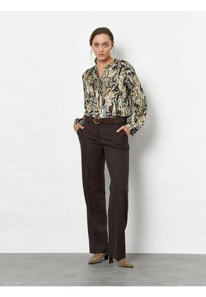 Donna in piedi con le mani in tasca, indossa una camicetta con stampa serpente, pantaloni marrone scuro, cintura marrone e stivaletti beige davanti a una parete bianca.