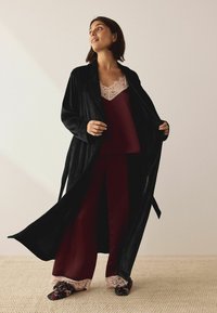 Schwarzes Samtgekleid, burgunderfarbener Satinoberteil mit Spitze, passende burgunderfarbene Hosen und floral gemusterte Hausschuhe auf einem strukturierten Teppich.