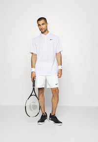 Balta polo marškinėliai su apykakle, juodas Nike logotipas. Balti šortai, juodi ir balti sportiniai batai. Laikantis teniso raketę, stovint ant vientiso fono.