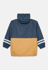 Wasserabweisende Jacke mit der oberen hälfte in Marineblau und der unteren hälfte in Senfgelb. Verfügt über elastische Bündchen und zwei weiße Streifen an den Ärmeln.