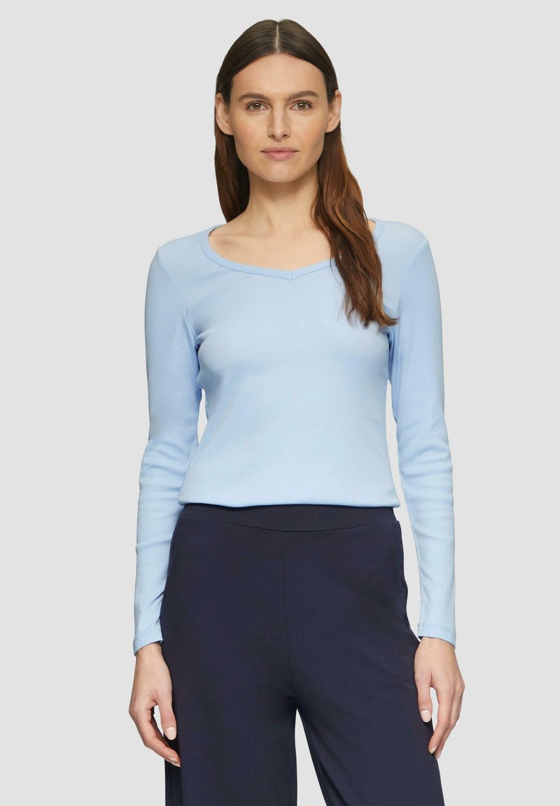 Hellblaues Langarmoberteil mit weitem V-Ausschnitt, figurbetontem Design und weicher Textur. Kombiniert mit dunklen Hosen. Neutrale Hintergrund.
