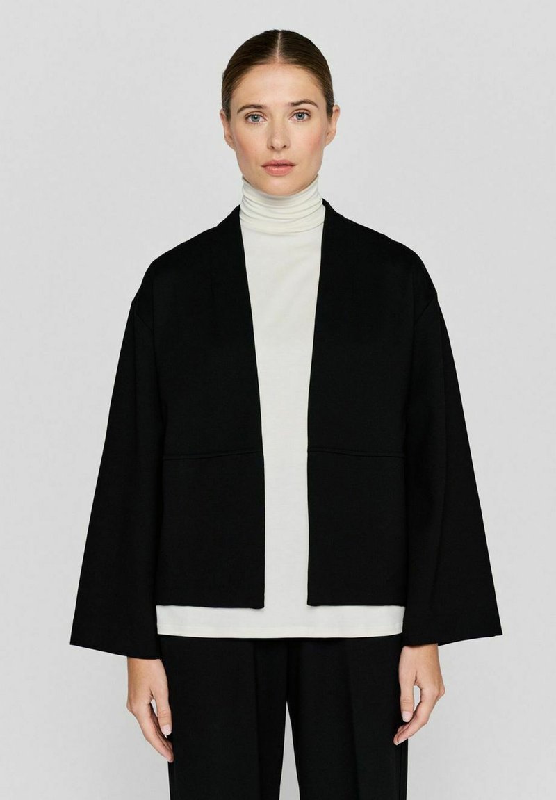 Zwarte kimono-stijl jas met brede mouwen, gemaakt van een gladde stof. Draag het over een lichtgekleurde coltrui. Eenvoudige, schone lijnen.
