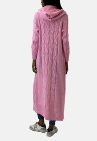 Długi różowy sweter z kapturem, wykonany z dzianiny warkoczowej, z prążkowanym wykończeniem na dole. Noszony na ciemnych dżinsach i jasnych butach.