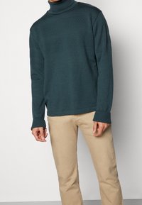 Dark teal turtleneck sweater with a textured pattern and ribbed cuffs, paired with light beige pants. Smooth fabric, casual fit.