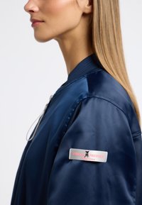 Marine satijnen bomberjack met geribde kraag, ritssluiting aan de voorkant en een logo patch op de mouw. Glad textuur en klassieke silhouet.