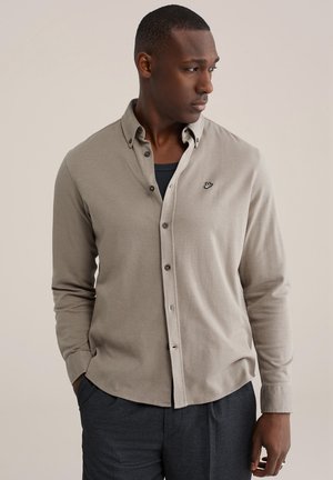 Man met een beige overhemd over een zwart T-shirt, donkere broek, kijkt naar rechts met handen in de zakken tegen een effen achtergrond.