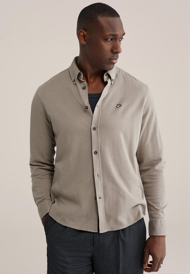 Uomo che indossa una camicia beige con bottoni sopra una maglietta nera, pantaloni scuri, guarda verso destra con le mani in tasca contro uno sfondo semplice.