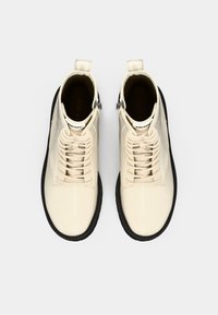 Botines de charol beige con suela de goma negra, punta redonda, cordones en la parte delantera y lengüetas en el talón. Diseño sencillo y contemporáneo.