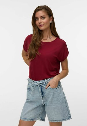 Femme aux longs cheveux bruns porte un t-shirt bordeaux et un short en jean bleu clair, debout avec une main dans la poche devant un fond uni.