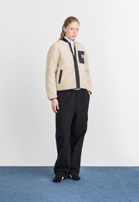 Beige fleece-takki mustilla yksityiskohdilla, vetoketjulla edessä ja sivutaskuilla, yhdistettynä mustiin cargo-housuihin ja mustiin kenkiin sinisellä lattialla.
