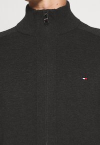 Ciemnoszary, dzianinowy sweter na zamek z wysokim kołnierzem i małym, trójkolorowym logo na lewej piersi. Prążkowana faktura i dopasowany krój.