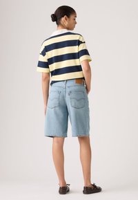 Γυναίκα με τα μαλλιά σε κότσο, φορώντας χρυσούς κρίκους σκουλαρίκια, πουκάμισο με ναυτικούς και κίτρινους ρίγες, ανοιχτόχρωμα τζιν σορτς μέχρι το γόνατο και σκούρα καφέ παπούτσια, στραμμένη πλάτη.