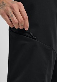Schwarze Sportpants mit einer Reißverschlusstasche. Der Stoff ist glatt mit einem dezenten Glanz, strukturiert für eine schmale Passform und betont die Funktionalität.