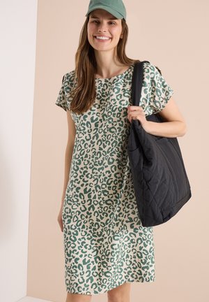 Lächelnde Frau mit grünem Leopardenmusterkleid und grüner Kappe, die eine große schwarze gesteppte Tasche auf der Schulter trägt.