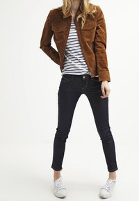 Chaqueta de ante marrón con diseño de cuello, dos bolsillos en el pecho, combinada con una camisa a rayas. Jeans oscuros y zapatillas blancas completan el conjunto.