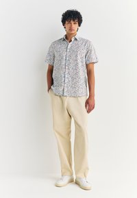 Chemise à manches courtes avec un motif floral multicolore ; pantalons beiges ; baskets blanches. Tissu lisse, coupe décontractée, design casual.