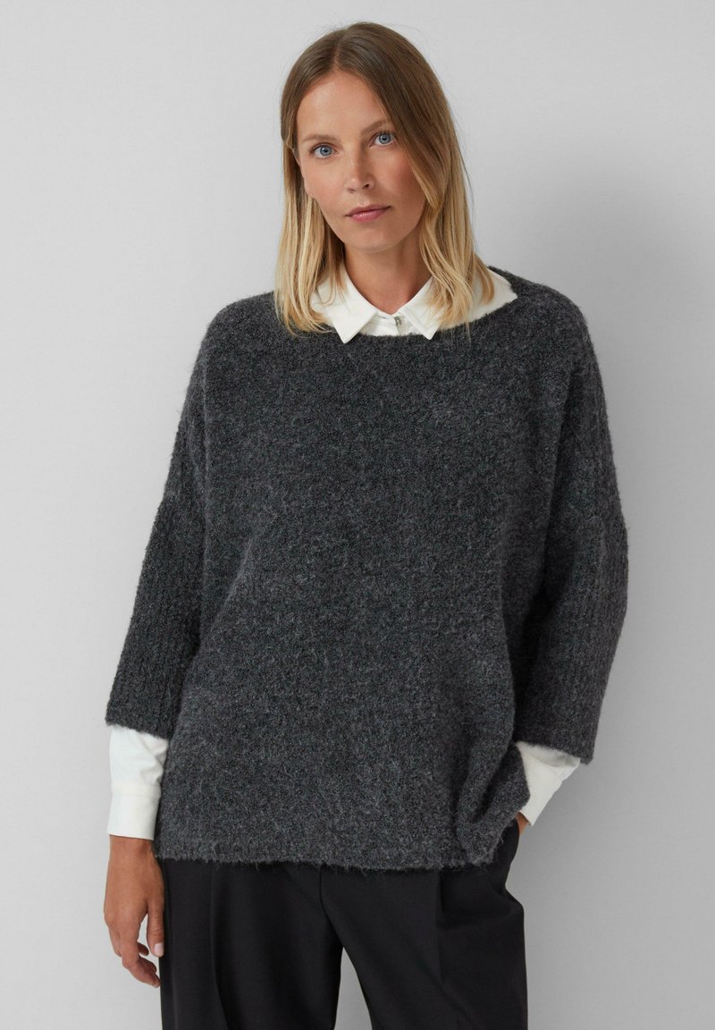 Femme aux cheveux blonds portant un pull en peluche gris charbon sur une chemise blanche à col et un pantalon noir, se tenant contre un mur gris clair.